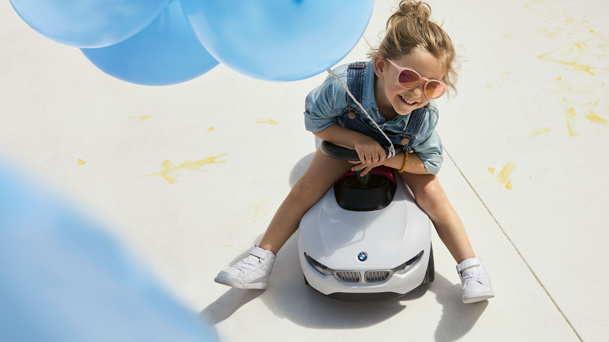 BMW Kids Kollekció. Egy rózsaszín napszemüveget viselő kislány boldogan ül a BMW Baby Raceren egy kék luftballon alatt.