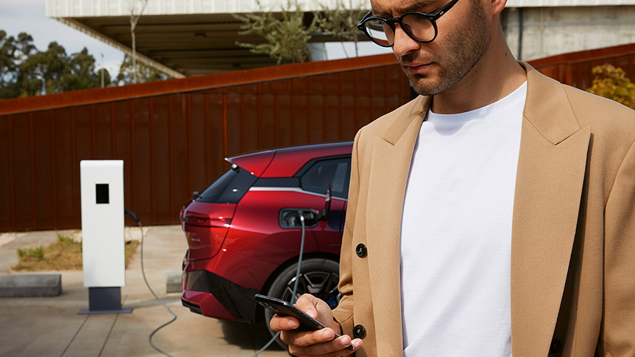 Egy okostelefont a kezében tartó férfi egy töltőállomáson álló BMW iX előtt Egy okostelefont a kezében tartó férfi egy töltőállomáson álló BMW iX előtt