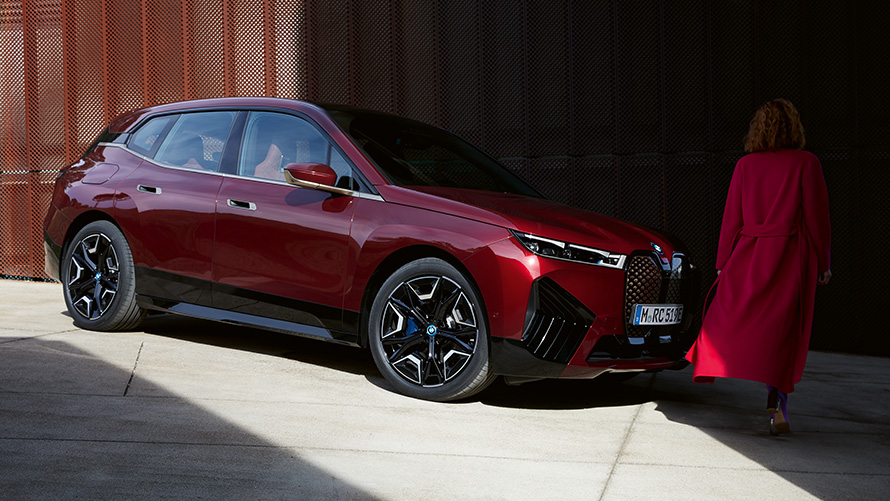 BMW Digitális kulcs Plus, BMW iX, egy rézsútos elölnézetből látható autó egy vörös kabátot viselő nővel BMW Digitális kulcs Plus, BMW iX, egy rézsútos elölnézetből látható autó egy vörös kabátot viselő nővel