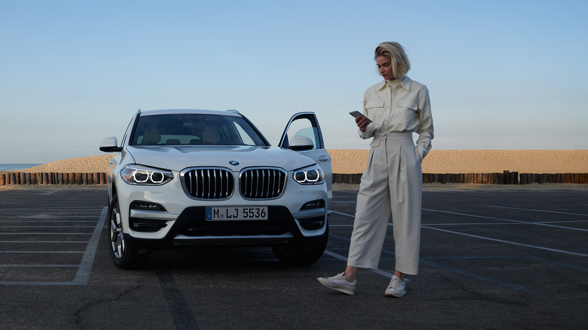 Egy nő a My BMW appot használja az okostelefonján Egy nő a BMW ConnectedDrive kínálatába tartozó My BMW appot használja az okostelefonján egy fehér BMW X5 előtt állva