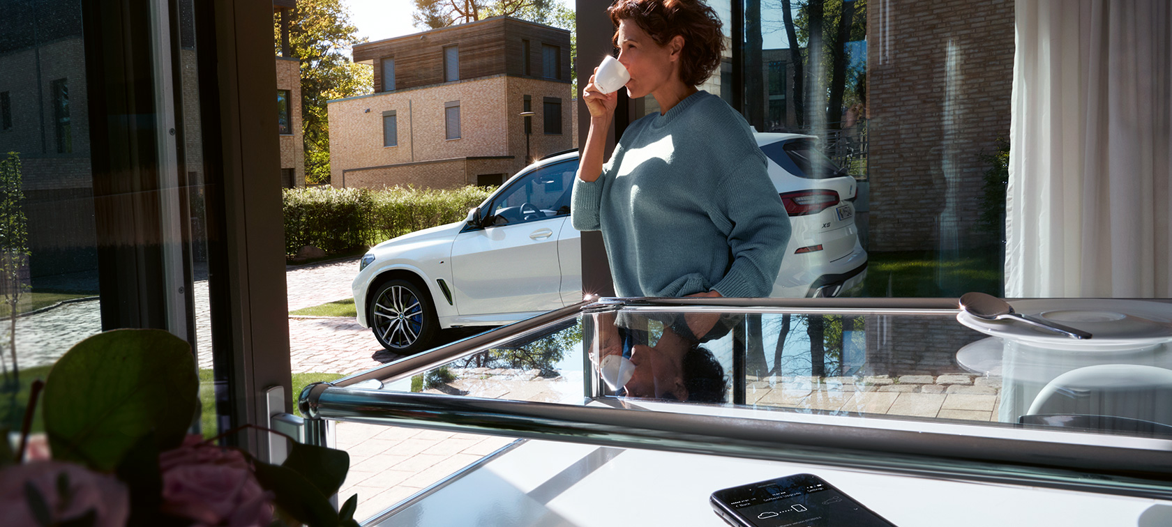 BMW távoli szoftverfrissítés Egy nő csészéből iszik, miközben a BMW Távoli szoftverfrissítés BMW X5-ösének szoftverét frissíti