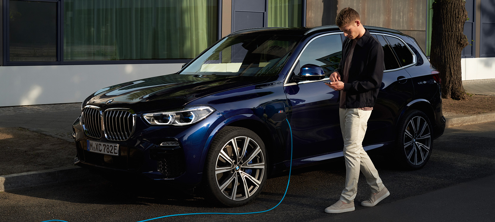 Egy férfi a My BMW appot használja az okostelefonján Egy férfi a BMW ConnectedDrive kínálatába tartozó My BMW appot használja az okostelefonján, miközben plug-in hibrid BMW X5-ösét tölti
