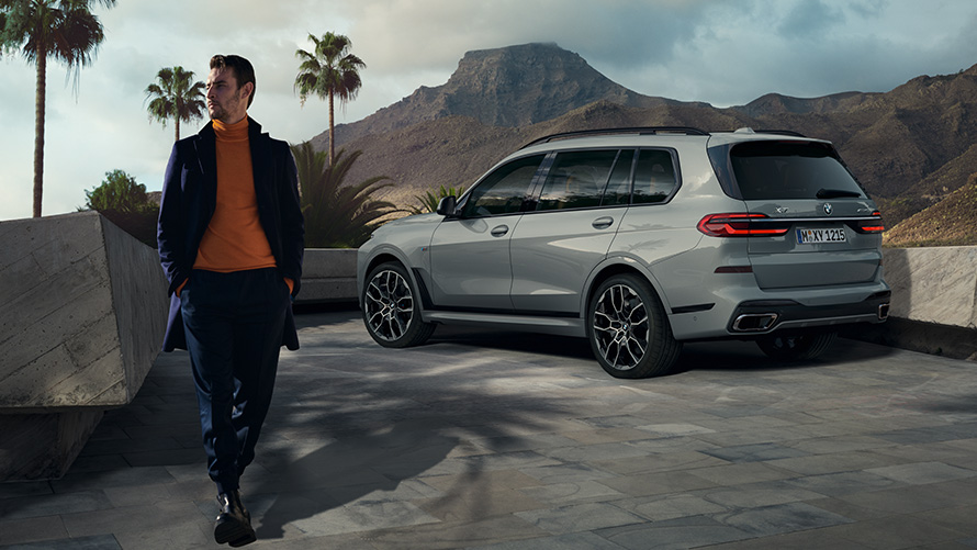 Egy parkoló BMW X7 (G07) előtt álló férfi, Távvezérlésű parkolás Távvezérlésű parkolás, egy pálmafák alatt parkoló, rézsútos hátulnézetből látható BMW X7 (G07) előtt álló férfi, a háttérben hegyi tájjal