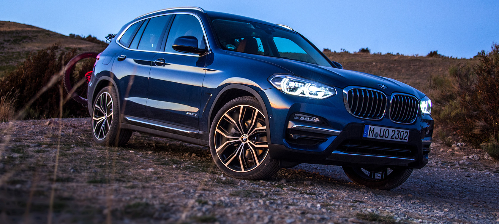 Egy sötétkék BMW X3 nyílt terepen Egy leparkolt, a hátsó tartóelemén mountain-bike-ot szállító, sötétkék BMW X3 nyílt terepen