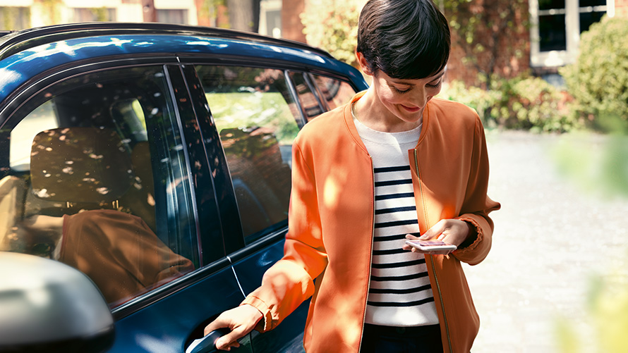 BMW Teleszerviz Egy okostelefont a kezében tartó nő áll BMW-je ajtajánál