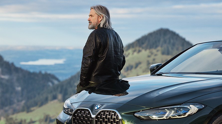 Egy férfi dől neki a BMW-jének, hegyvidékkel a háttérben BMW tavaszi-nyári tartozékok, egy férfi dől neki a BMW-jének, hegyvidékkel a háttérben