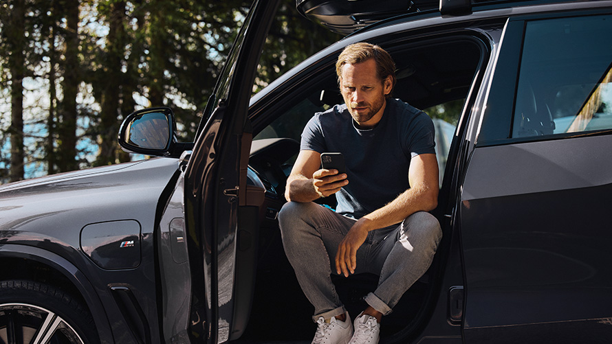 Egy, a BMW-jében ülő férfi az okostelefonját nézi BMW tavaszi-nyári tartozékok, egy, a BMW-jében ülő férfi az okostelefonját nézi