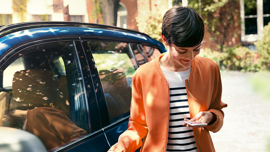 BMW nyári ajánlatok, BMW Teleszerviz BMW nyári ajánlatok, BMW Teleszerviz, egy, az okostelefonját használó nő áll a BMW-je előtt