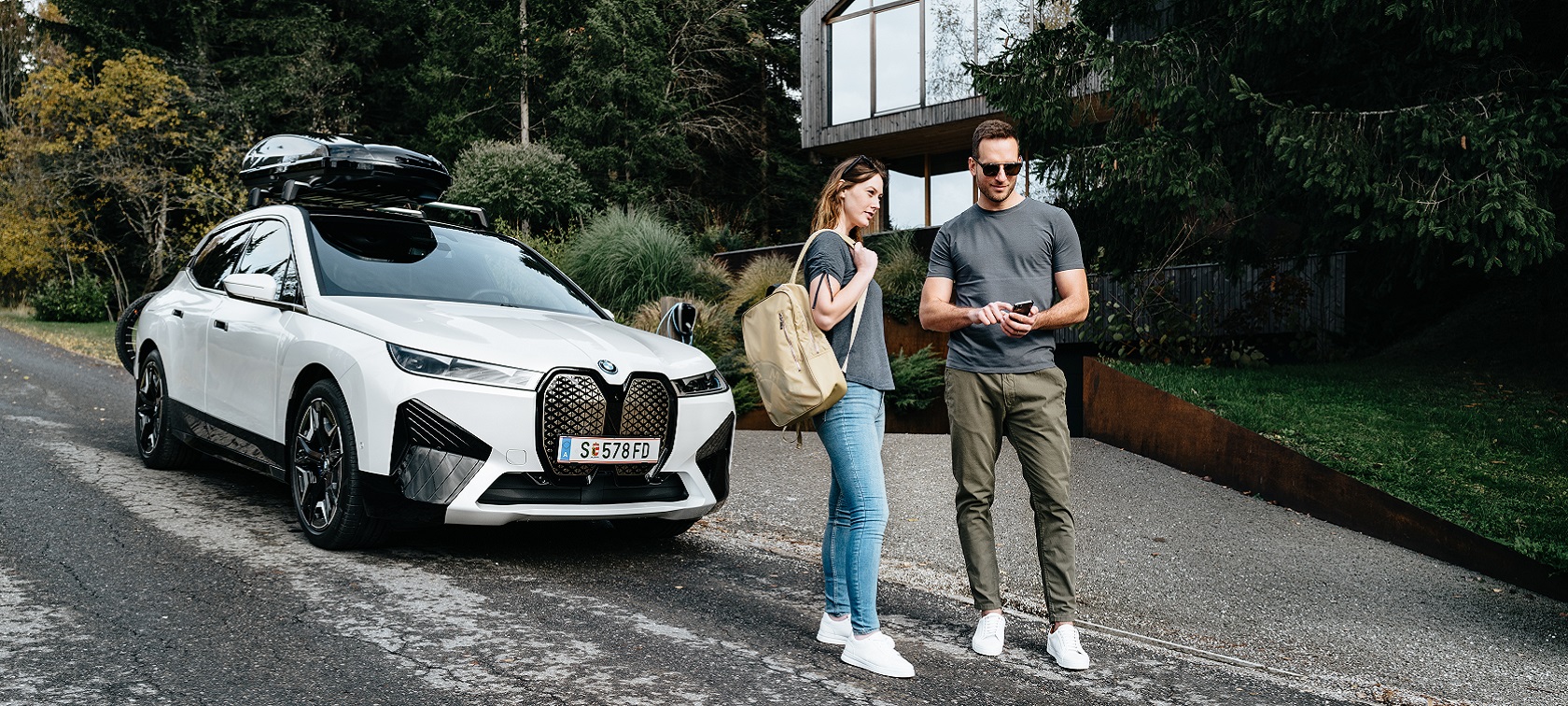 Egy, a kezében telefont tartó férfi és egy hátizsákos nő áll egy BMW iX előtt. Egy, a kezében telefont tartó férfi és egy hátizsákos nő áll egy BMW iX előtt.