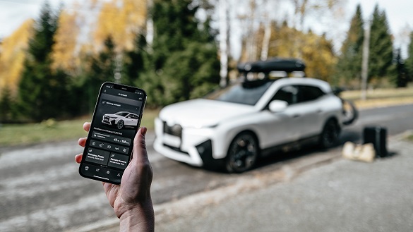 Egy telefon, a kijelzőjén a My BMW app felületével, a háttérben egy tartozékokkal ellátott BMW iX. Egy telefon, a kijelzőjén a My BMW app felületével, a háttérben egy tartozékokkal ellátott BMW iX.