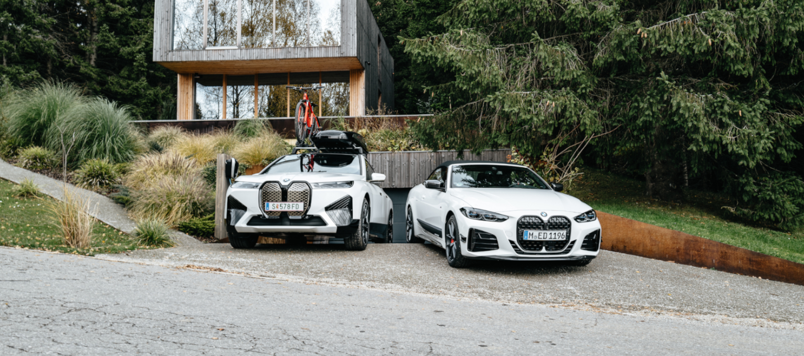 Egy modern épület előtt álló, tartozékokkal ellátott BMW iX és egy BMW M440i Egy modern épület előtt álló, tartozékokkal ellátott BMW iX és egy BMW M440i
