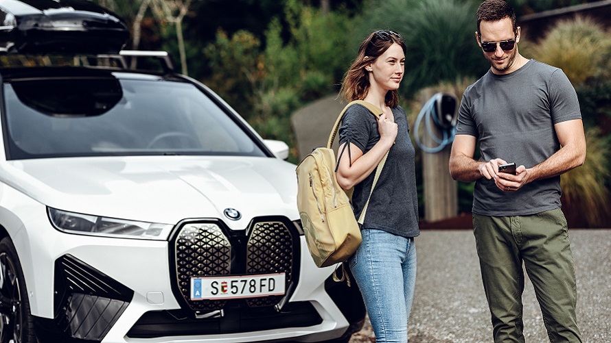 Egy, a kezében telefont tartó férfi és egy hátizsákos nő áll egy BMW iX előtt. Egy, a kezében telefont tartó férfi és egy hátizsákos nő áll egy BMW iX előtt.
