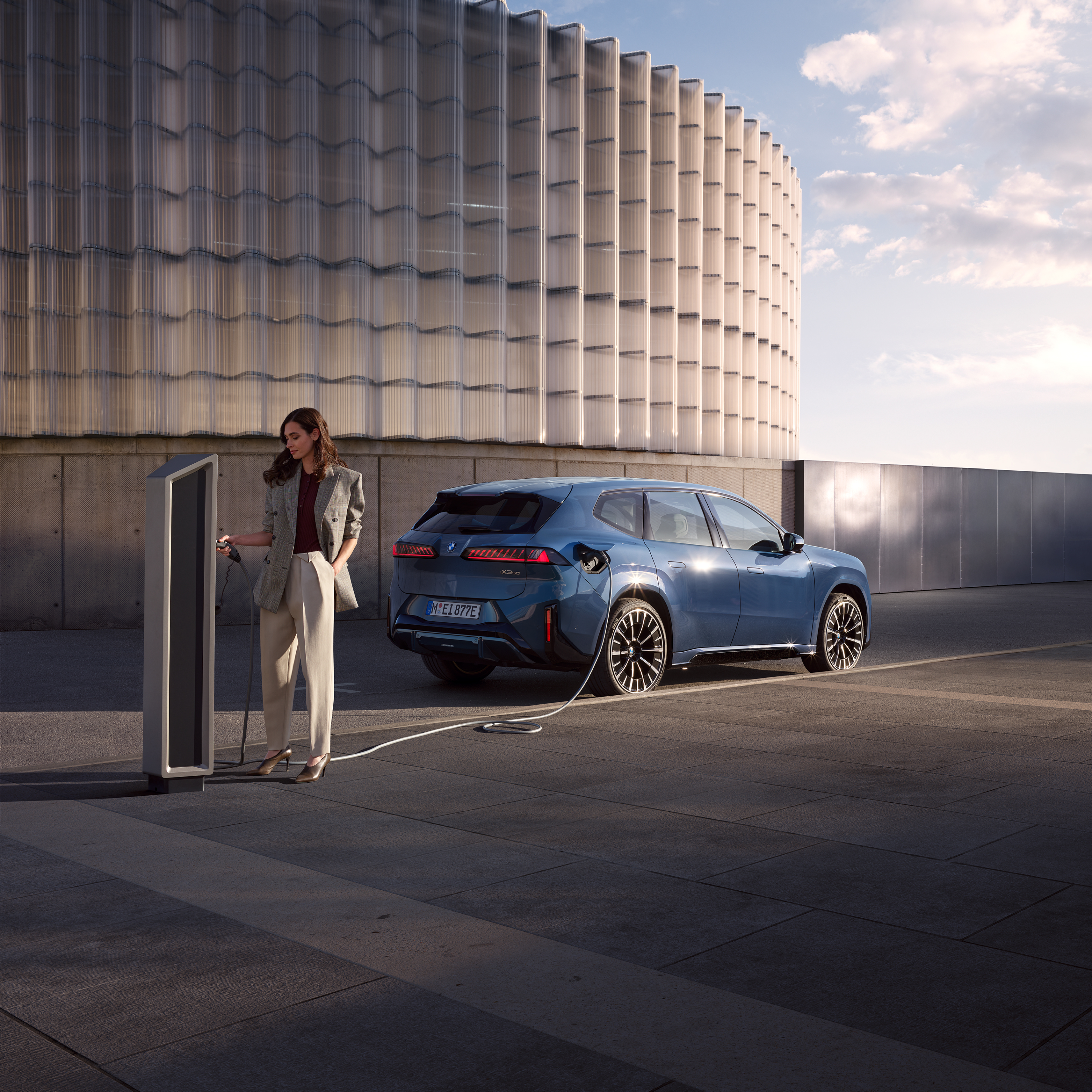 Egy nő áll egy nyilvános töltőállomáshoz csatlakoztatott BMW iX3 mellett